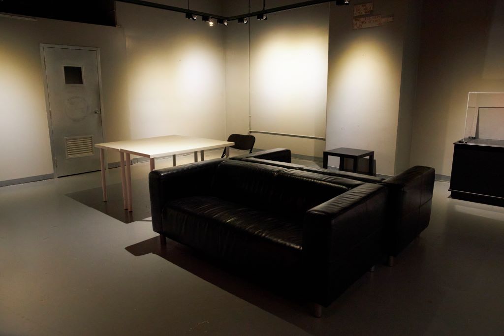 Dimly lit room with a black sofa and tables against beige walls.