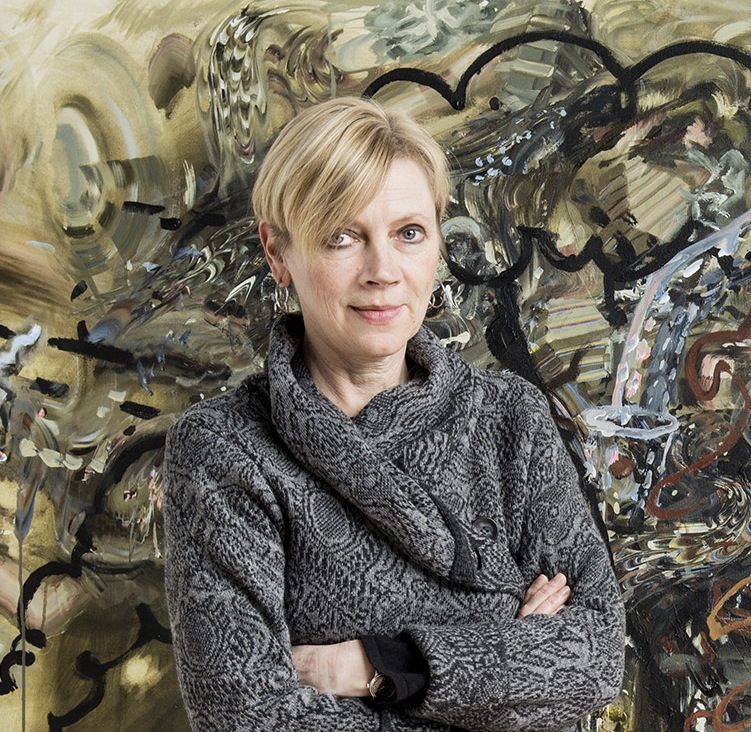 Confident woman with arms crossed in a textured gray sweater stands before a complex mechanical backdrop.