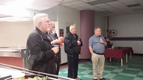 A group of older men standing indoors, attentively listening.