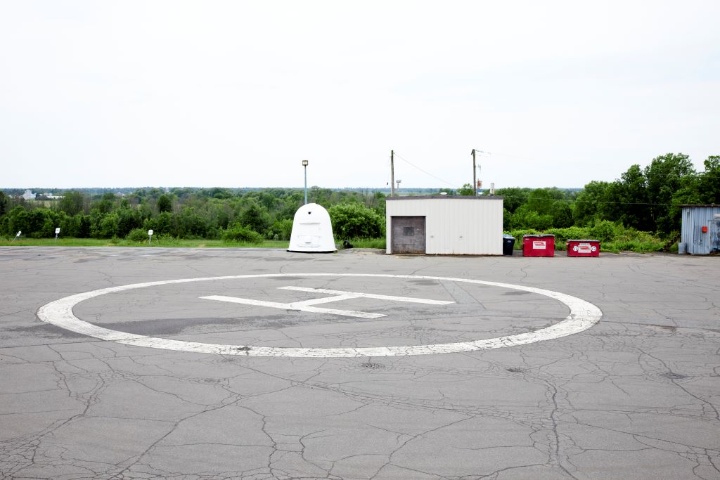 A helipad with a white H marking, surrounded by greenery and small structures.