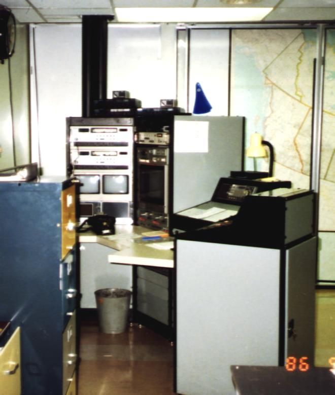 Old computer equipment and electronics in a cluttered room with a map on the wall.