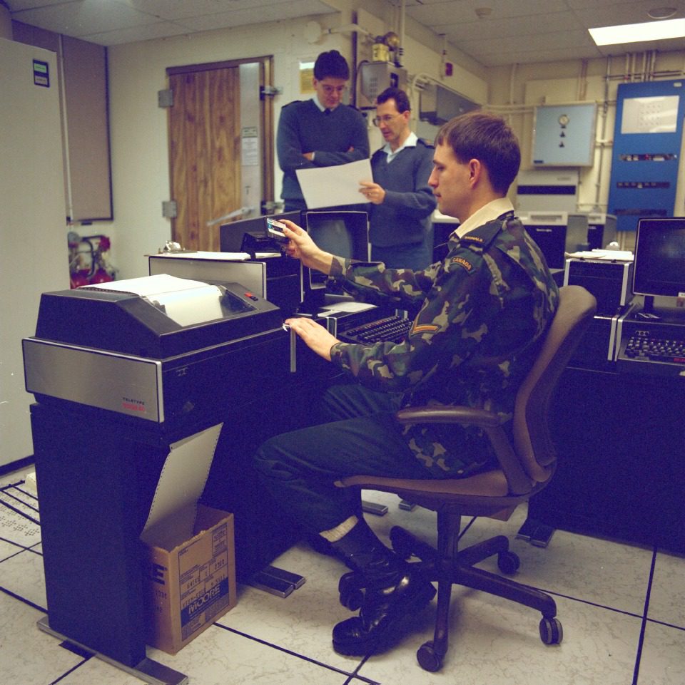 Two men operating vintage computer equipment in a control room.