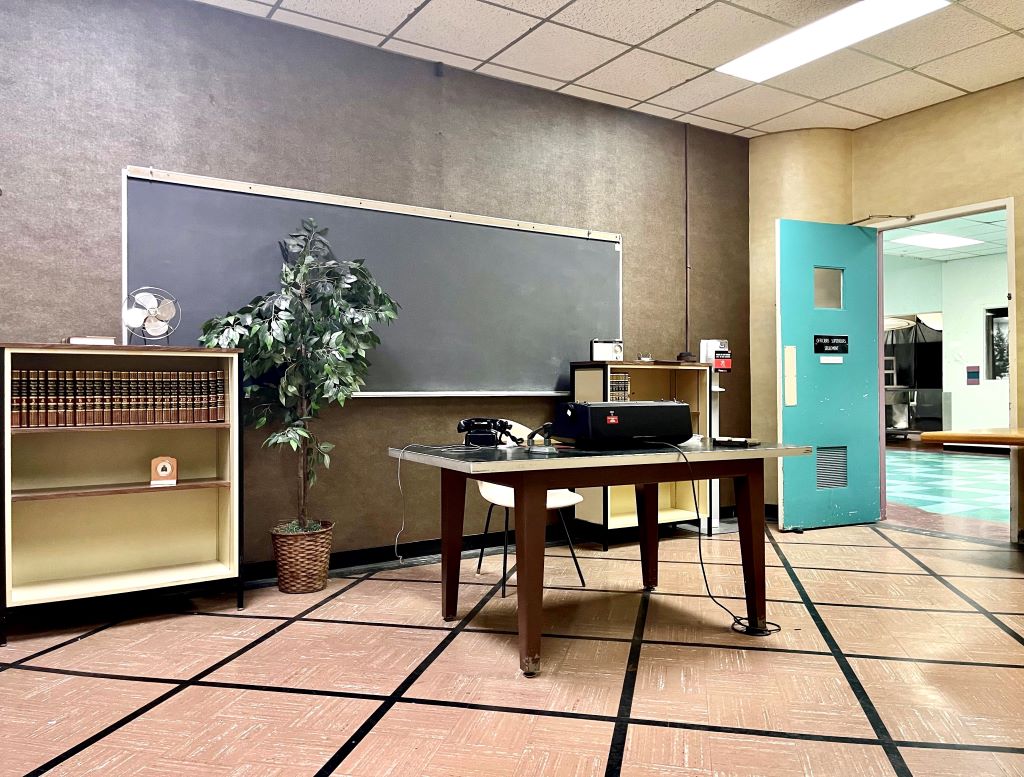 A classroom with a chalkboard, teacher's desk, and potted plant.