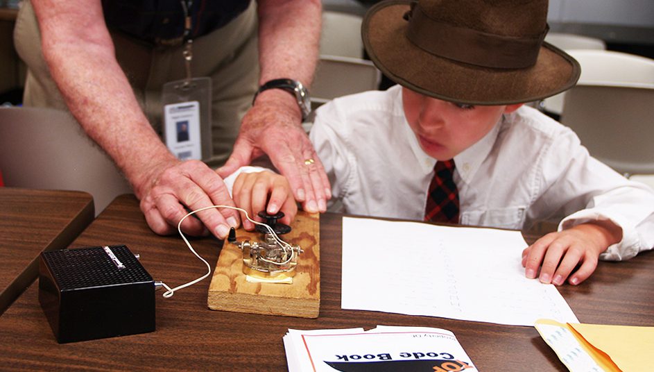 Boy learning Morse code with a wooden telegraph key and instructor's hands guiding him.