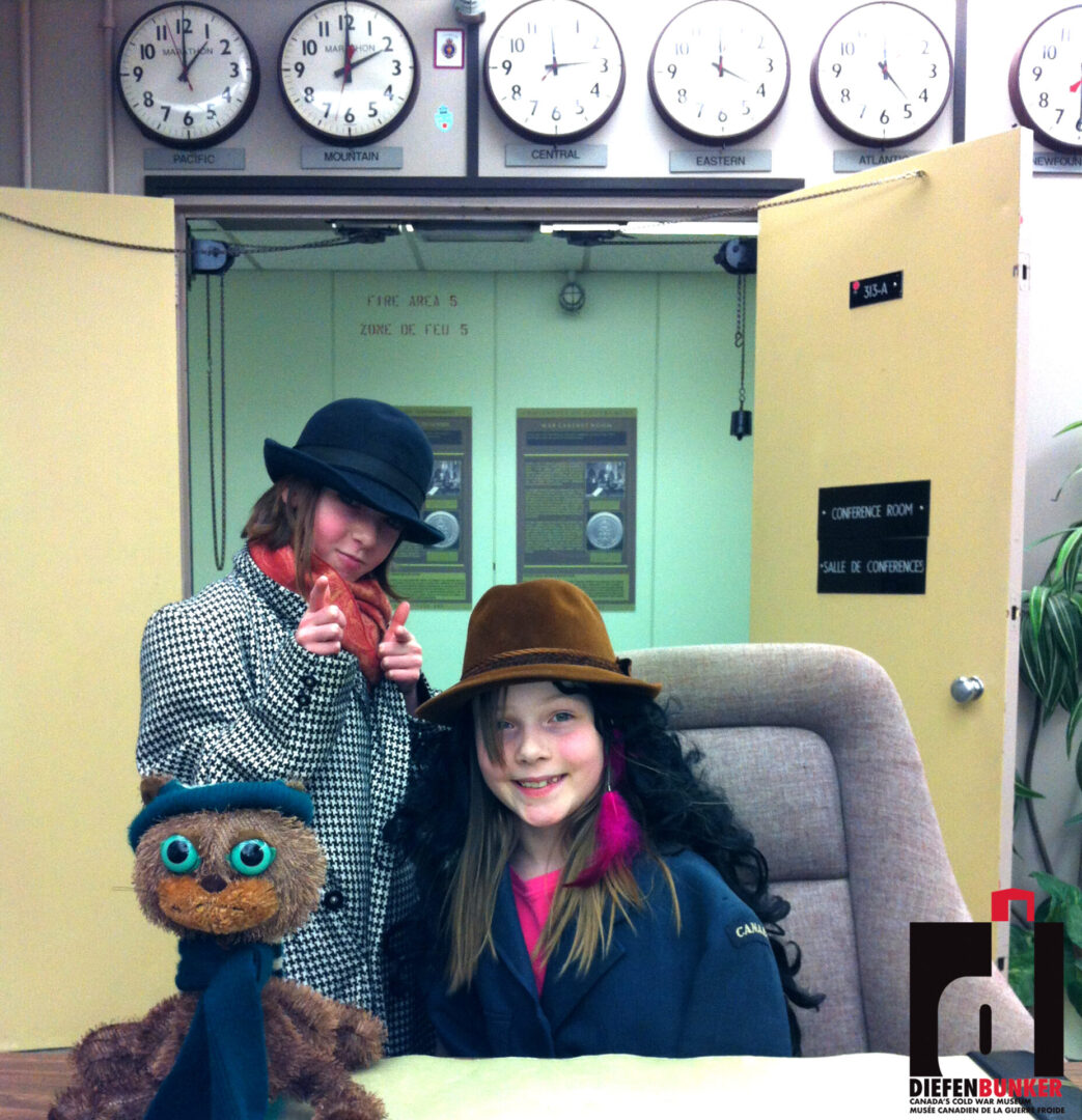 Two people wearing hats posing indoors with clocks on the wall behind them.
