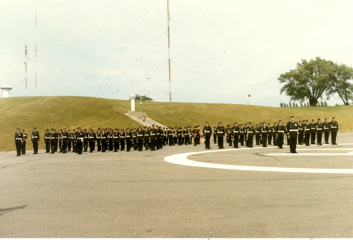 Military personnel standing in formation on a parade ground with a monument in the background.