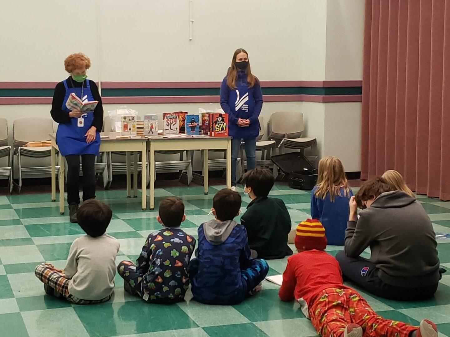 Two adults reading to a group of children seated on the floor in a library.
