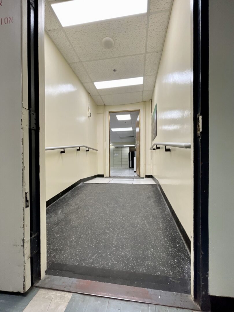 A narrow hallway with white walls and a gray carpet leading to a door.