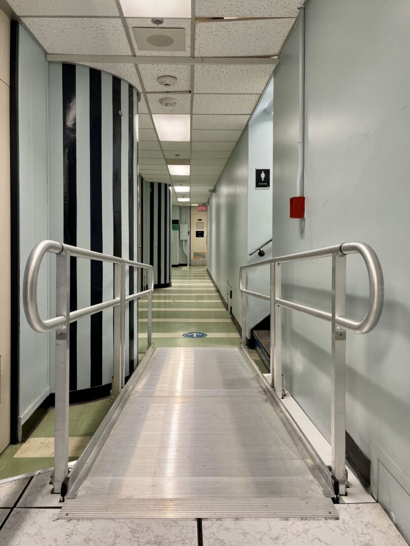 A wheelchair-accessible ramp inside a building with handrails and a fire alarm on the wall.