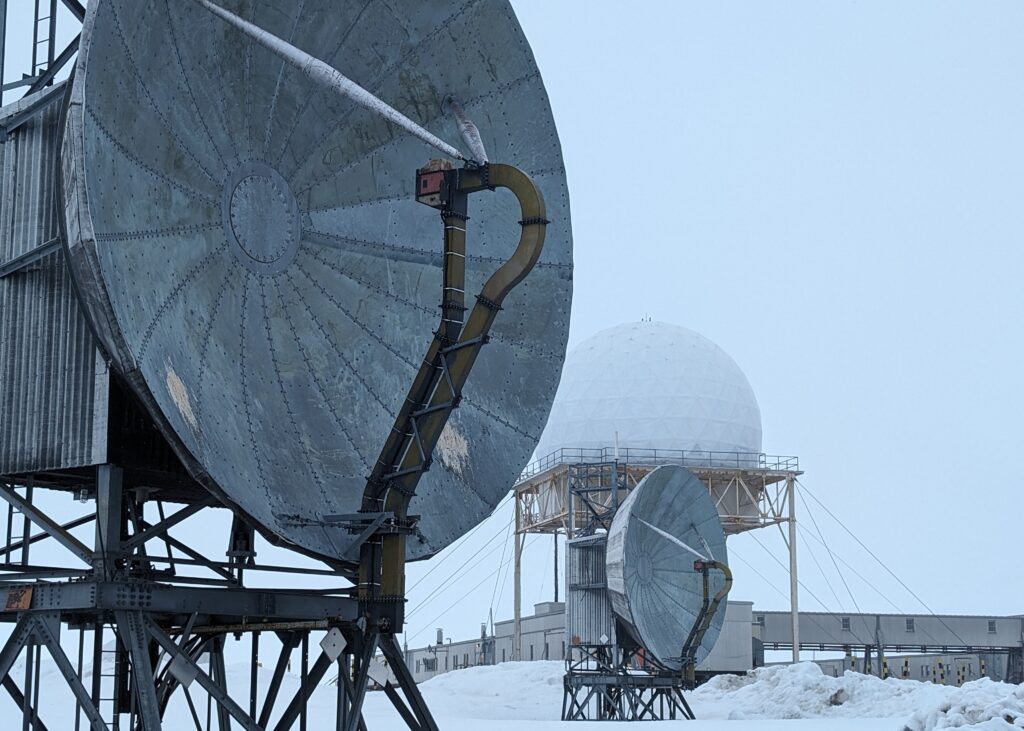 Two large grey parabolic antennae set in a snow-covered area with a large white dome behind them.