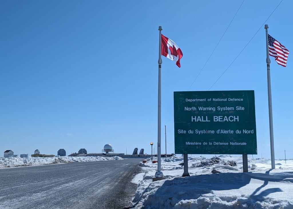 A green sign with the text "Hall Beach" in between a Canadian and American flag, beside a road in a snow-covered area.