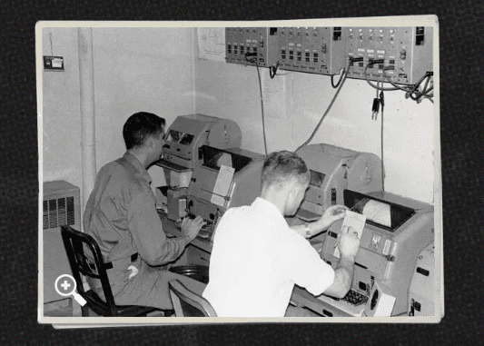 Black and white photo of two men sit in front of Cold War era machinery.