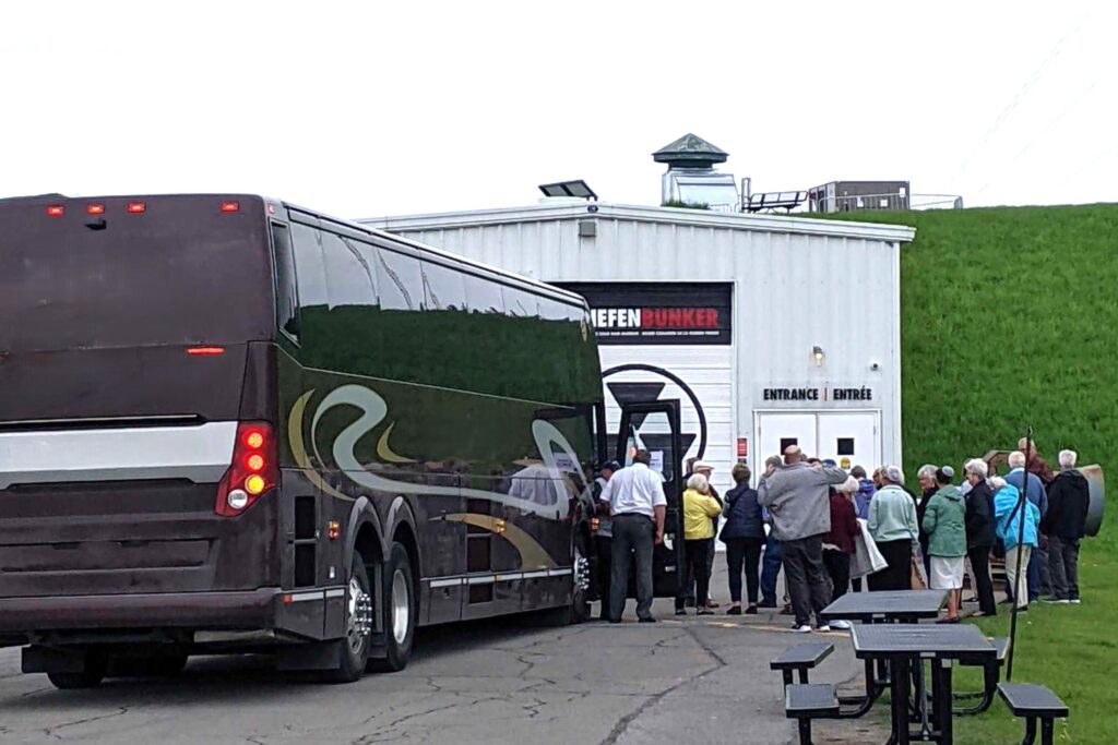 Group of adults disembarks a tour bus parked in front of a white building.