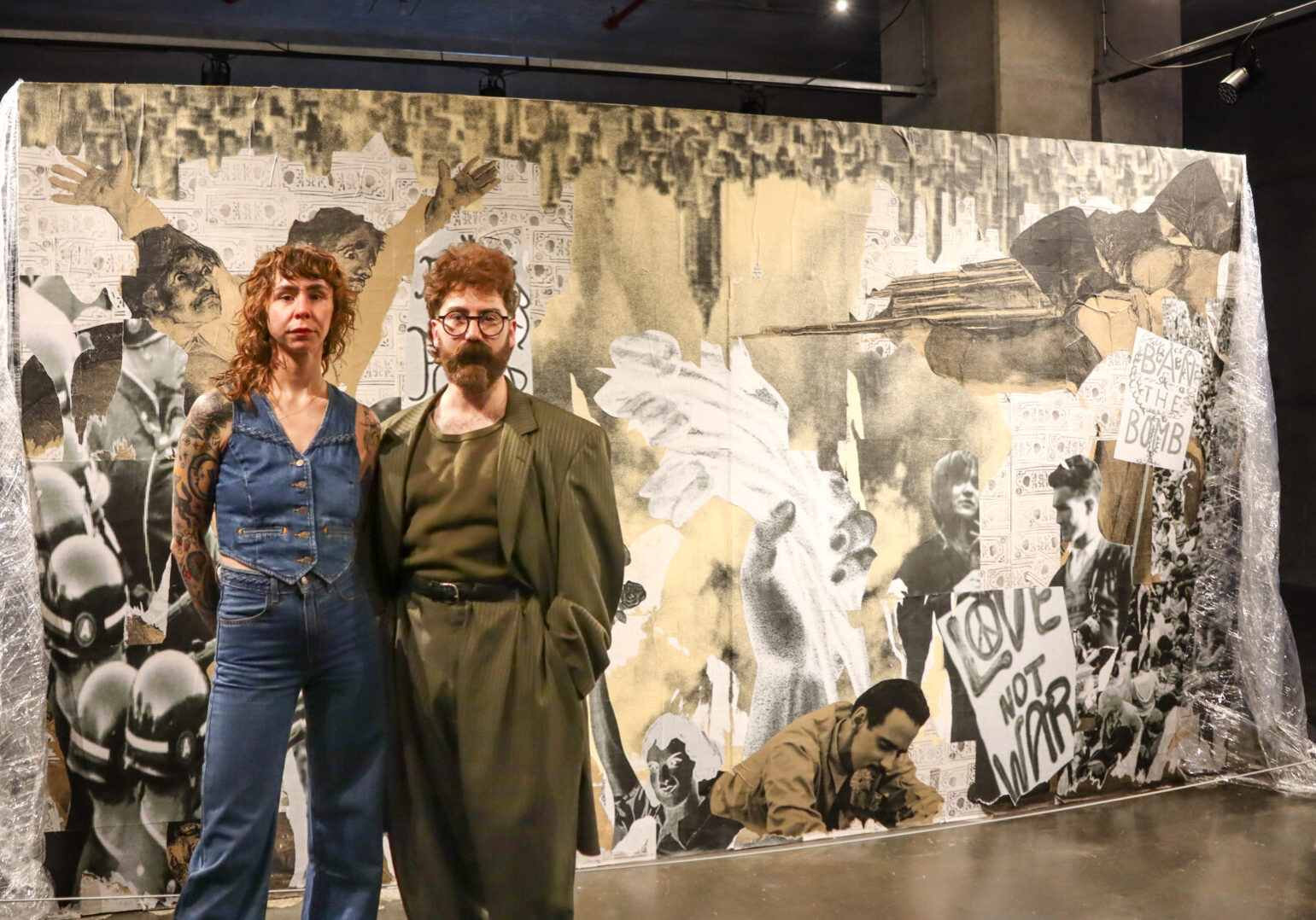 Diefenbunker 2025 Artists-in-Residence Olivia Johnston and Neeko Paluzzi stand in front of one installation in their exhibition, a large wall structure with collaged imagery from Cold War protests.