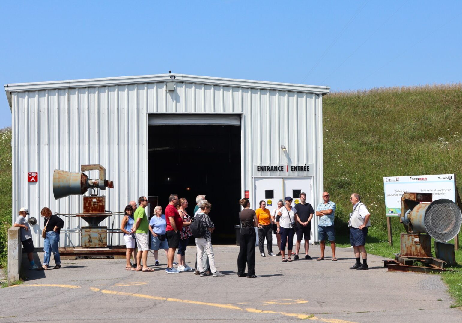 Group of people gathered outside a metal building on a sunny day.